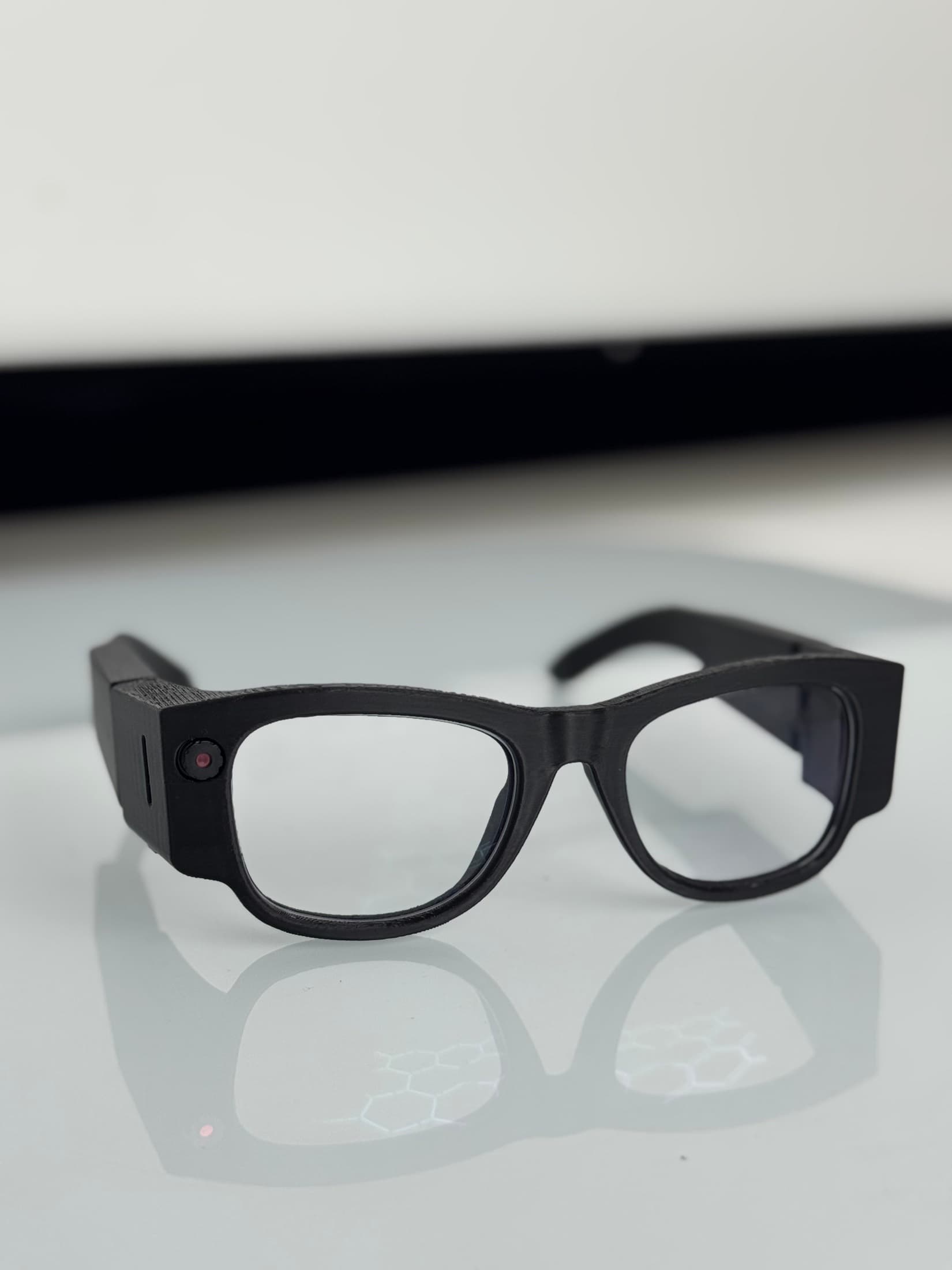 The EchoPath glasses from the front on a white desk. Matte black frame, thick temples, a small camera and indicator on the left temple block.